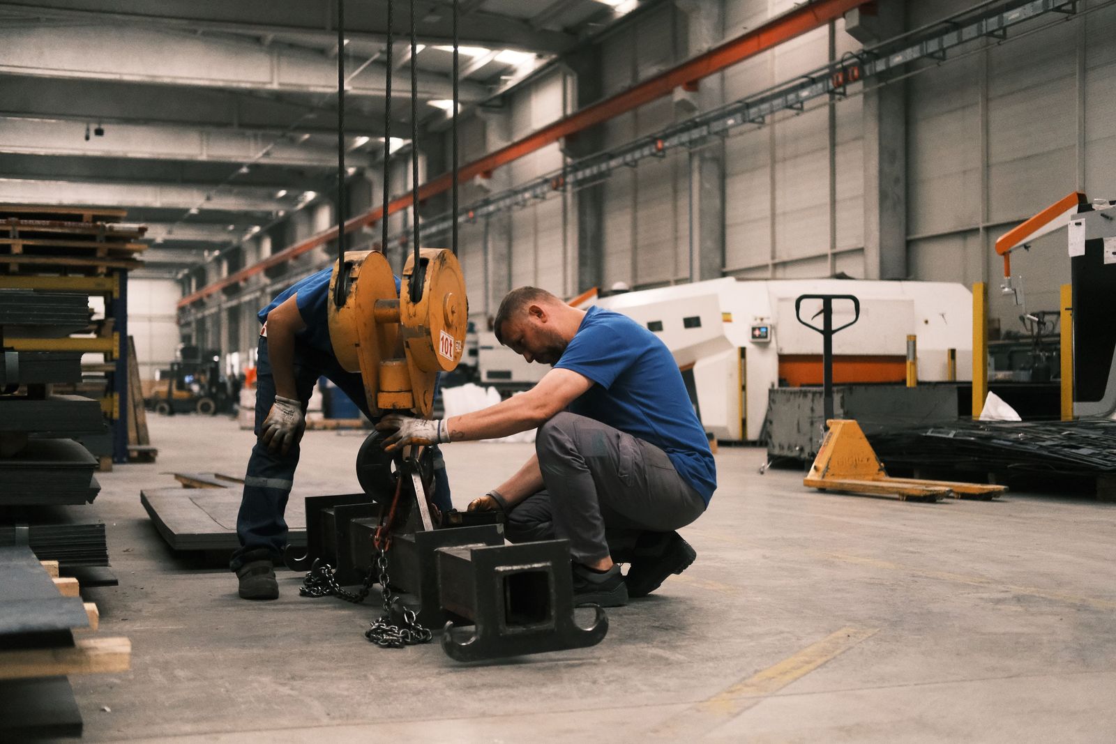 Two workers rigging a heavy machine for a crane lift in a factory hall.