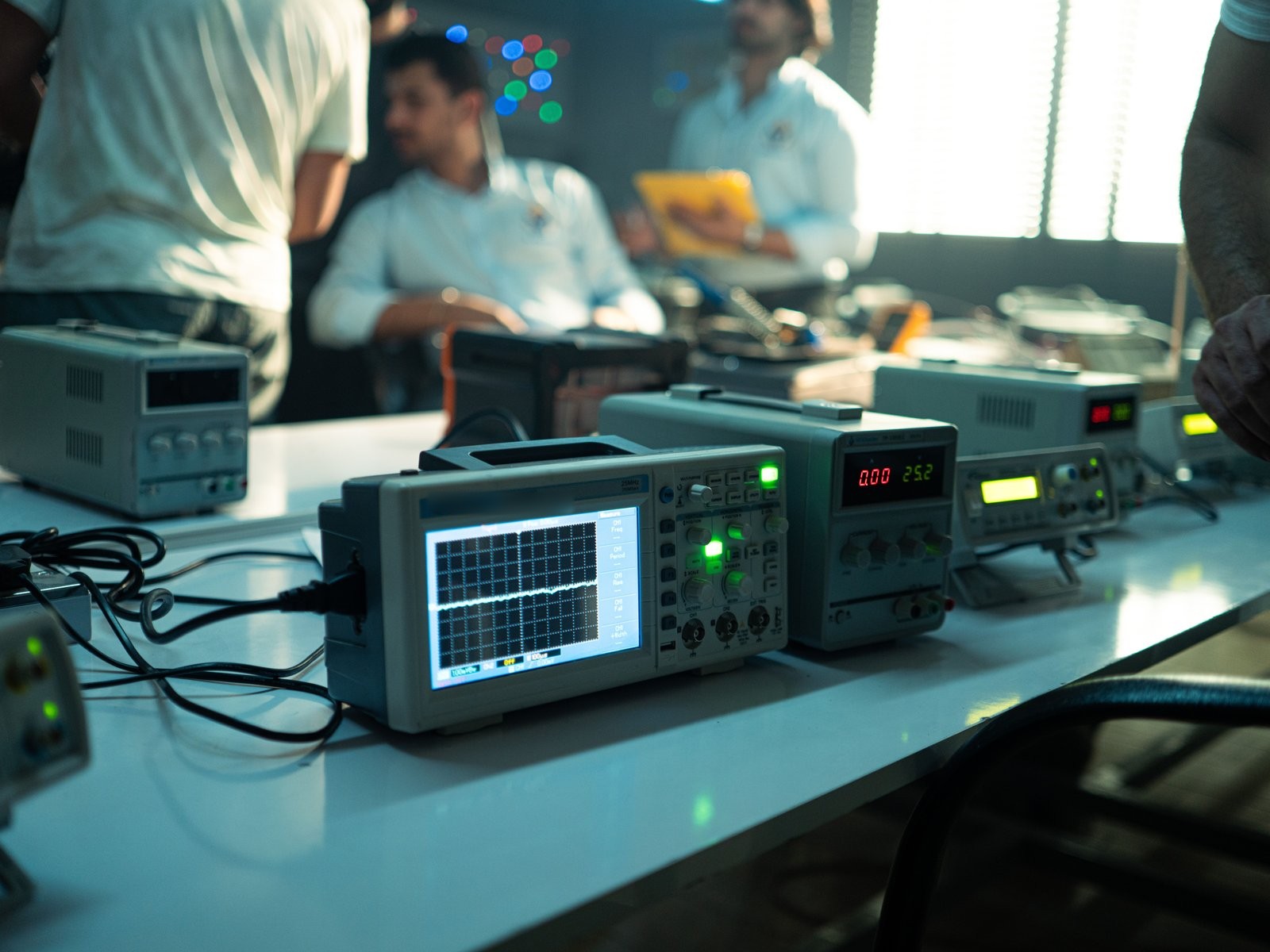 Oscilloscope and power supplies on a test bench, engineers working behind.